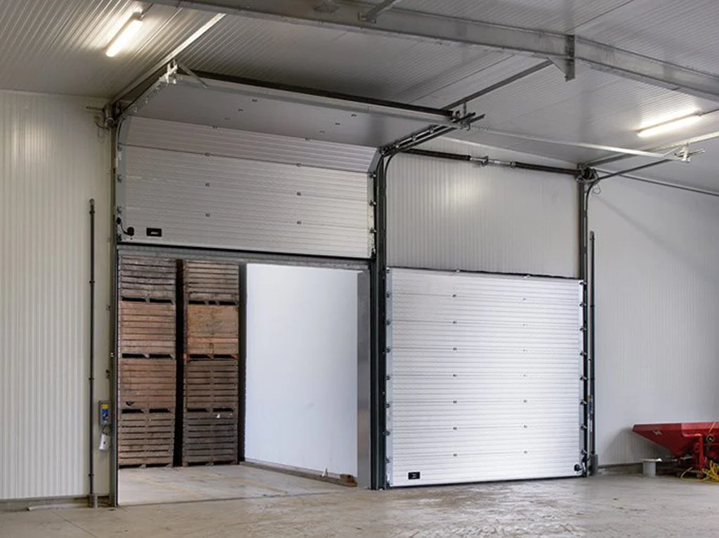 Two white insulated sectional overhead doors in a warehouse, one raised over stacked crates